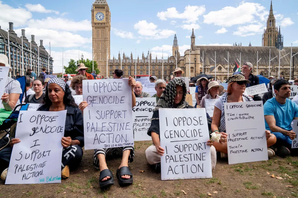 photo of peaceful protesters, in London, sitting on the ground and holding signs in support of 'Palestine Action" and opposing genocide 
(NATASHA QUARMBY/ALAMY)