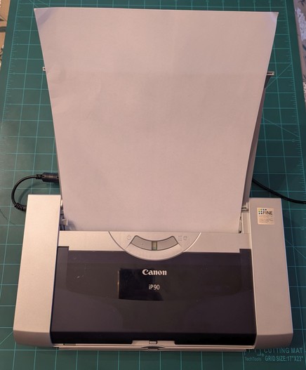 A Cannon iP90 inkjet printer. The lid is opened up and and a sheet of paper is loaded.