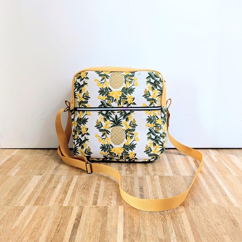 Handbag with a flower-pineapple pattern, the pineapples are golden. It has ha square pattern with a zipper on top and roughly at golden cut in the front.