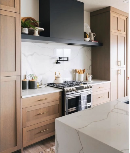 A high-end kitchen with 10’ ceilings. White oak full length cabinets flank a slide-in range. White engineered stone quartz on the countertops and backsplash, with a sleek matte black vent hood above. The sides extend to form shelves on either side, that end at the full-length cabinets. An island with waterfall edge in the same quartz can be seen in the foreground.