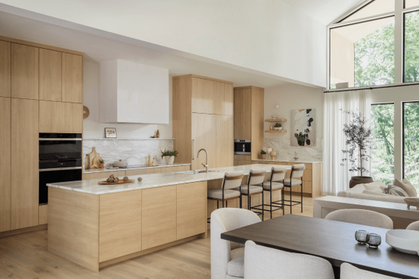 A contemporary kitchen in a high-end home. Flat front white oak cabinets, white stone countertops, and a rectangular white hood vent over an induction cooktop. A giant island has space for four countertop-height chairs AND about 9 ft of pure uninterrupted counter space next to it.