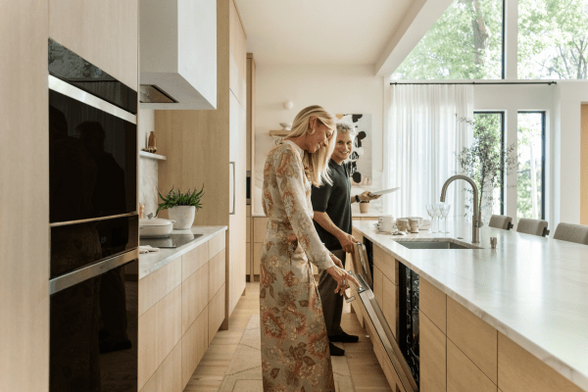 A contemporary kitchen in a high-end home. Flat front white oak cabinets, white stone countertops, and a rectangular white hood vent over an induction cooktop. A tall beautiful blonde woman wearing a long-sleeved light floral pattern dress smiles as she opens the integrated dishwasher installed in the island opposite the cooktop wall. A middle-aged smiling white man wearing a dark top and trousers stands about 4 feet behind her opening a second dishwasher. These people have a lot of dishes to wash, but they’re super happy about it, probably because they have a beautiful kitchen.