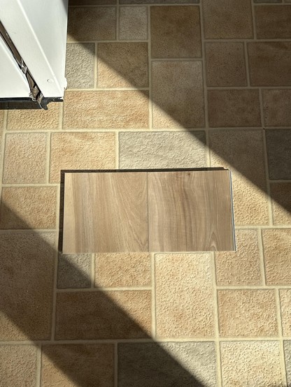 Two samples were f Lifeproof Ledge Point Pecan next to each other, on a sheet vinyl floor that consists of beige and grey faux stones. The LVP pecan samples are a medium brown sandy beige color, with light and dark streaks in the pecan grain. They fall within a big streak of morning sunlight. The corner of a white fridge peaks out of the top left corner.